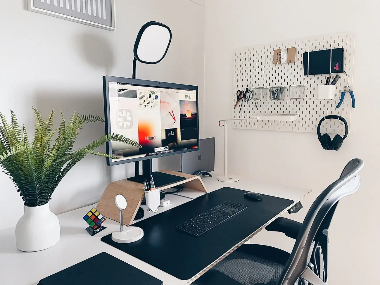 Moderner Schreibtisch mit Monitor, Tastatur, Maus, Lampe, Pflanze und Zubehör an einer Lochwand.